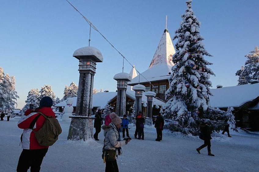 A snowy landscape with a large Christmas tree, a lodge, and people walking. 1