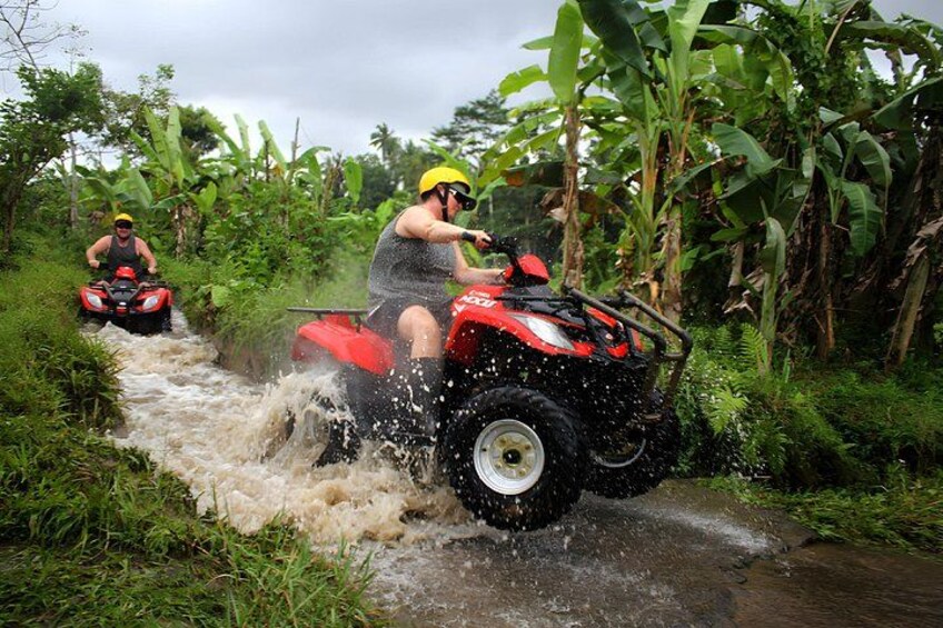 Two people riding red all-terrain vehicles through a shallow water crossing surrounded by lush greenery. 2