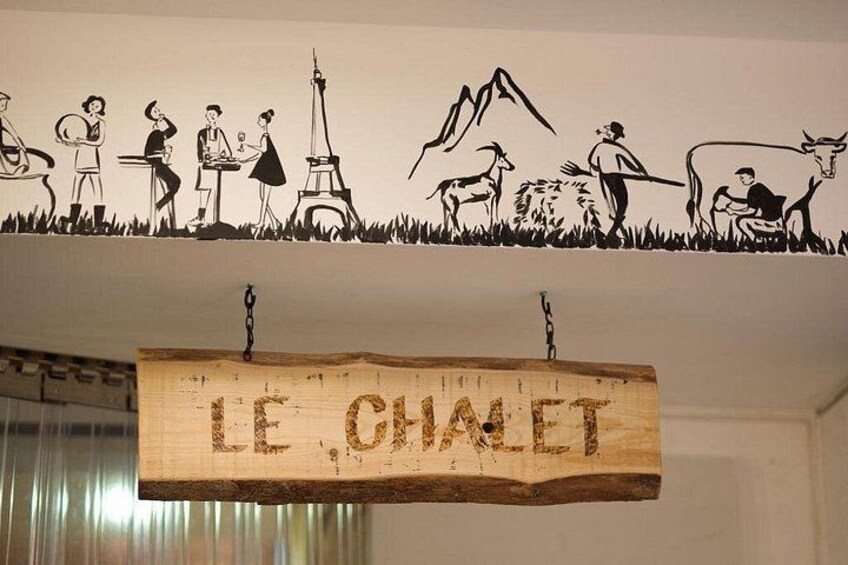 Une enseigne en bois portant l’inscription « LE CHALET » est suspendue au plafond, et une illustration en noir et blanc représentant diverses scènes se trouve au-dessus. 3