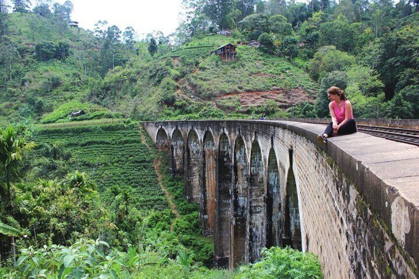 A woman sitting on a bridge with arched railings overlooking a lush green tea plantation. 4