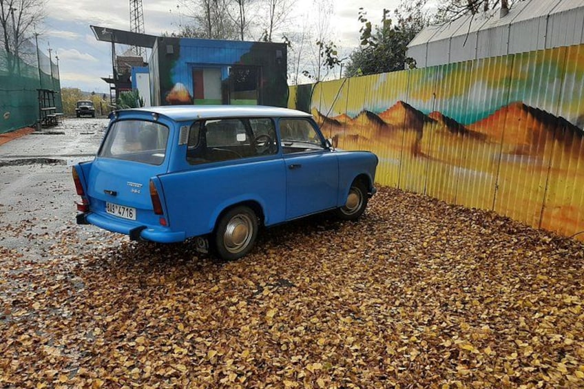 En blå bil parkeret på et løvbelagt område med et farverigt murmaleri i baggrunden. 2