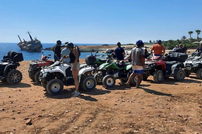 Un groupe de personnes se tient près de quelques quads sur une plage, avec l’épave d’un navire en arrière-plan. 1