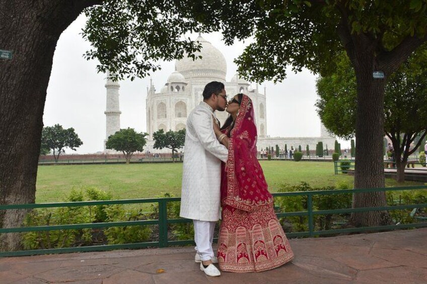 A couple embracing in front of the Taj Mahal, with a tree and a green railing in the foreground. 3