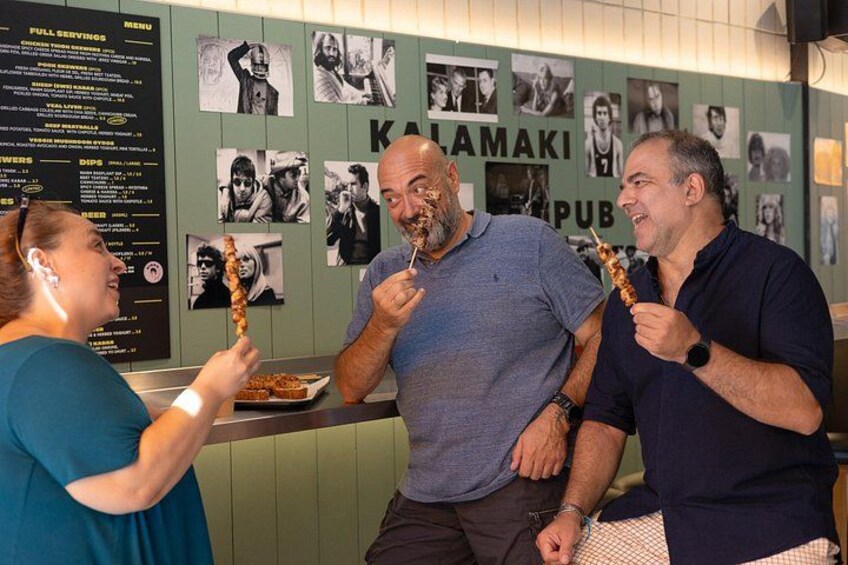 Three people eating skewers at a restaurant with a menu board and wall photos. 2
