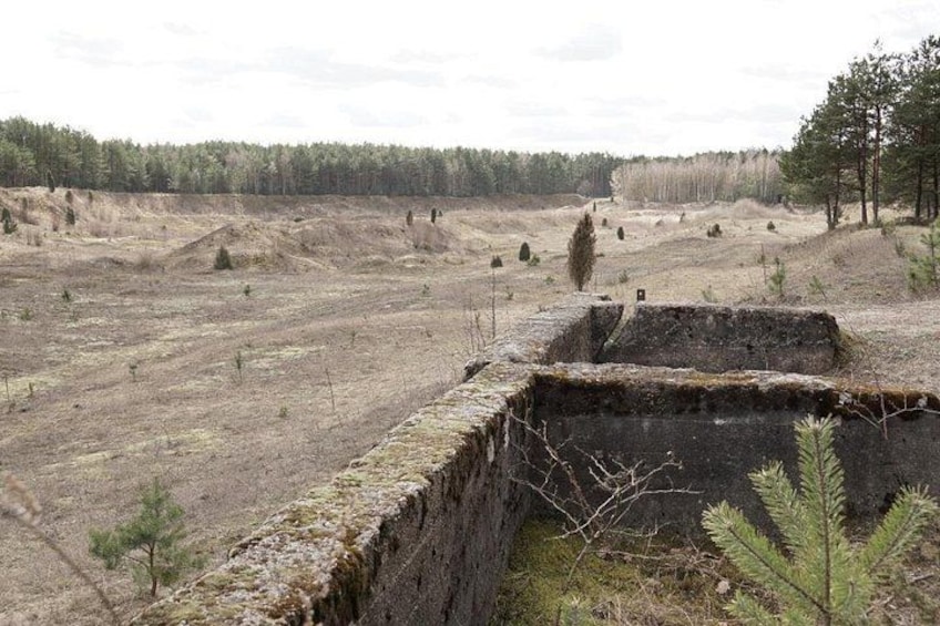 Eine karge Landschaft mit einer großen Betonkonstruktion, spärlicher Vegetation und einem Wald im Hintergrund. 5
