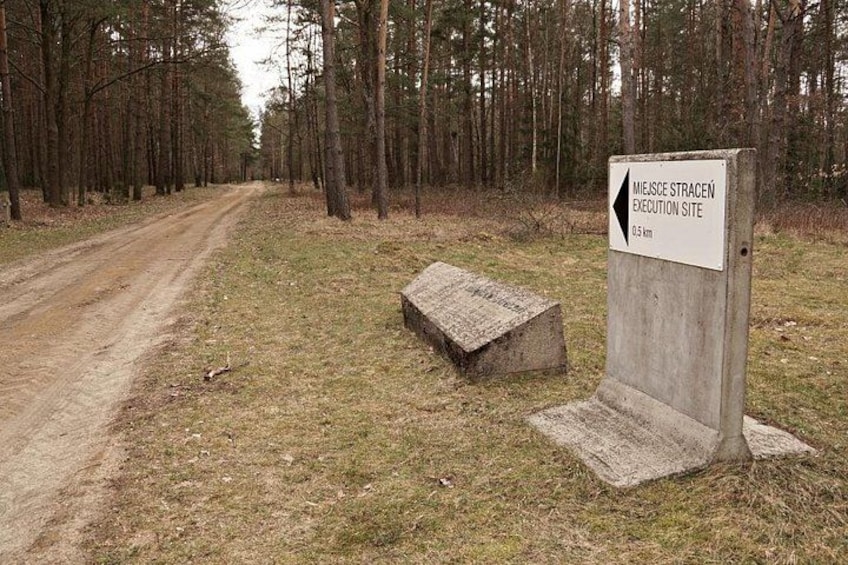 Ein Feldweg führt in einen Wald, ein Schild weist auf die Hinrichtungsstätte Miejsce Stracene und eine Betonkonstruktion hin. 4