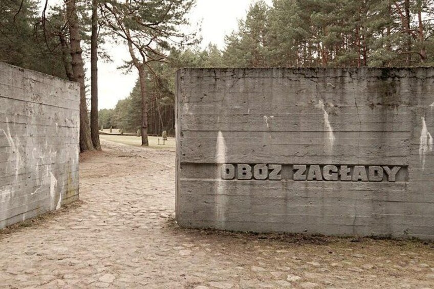 Eine Betonwand mit der Aufschrift 'OBOZ ZAGADY' und ein Pflasterweg, der durch ein bewaldetes Gebiet führt. 3