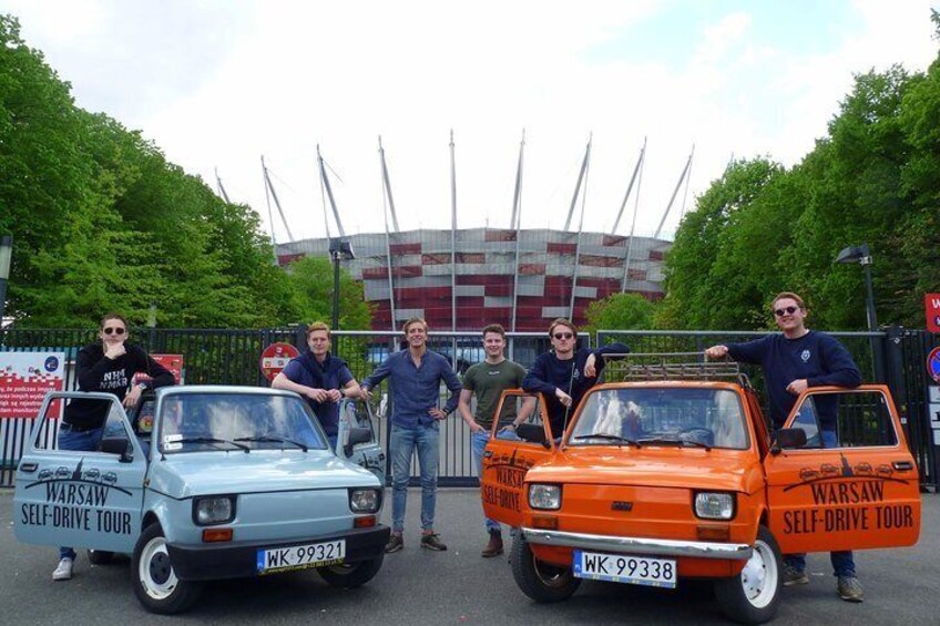 Two vintage cars with 'Warsaw Self-Drive Tour' written on them, parked in front of a stadium. 2