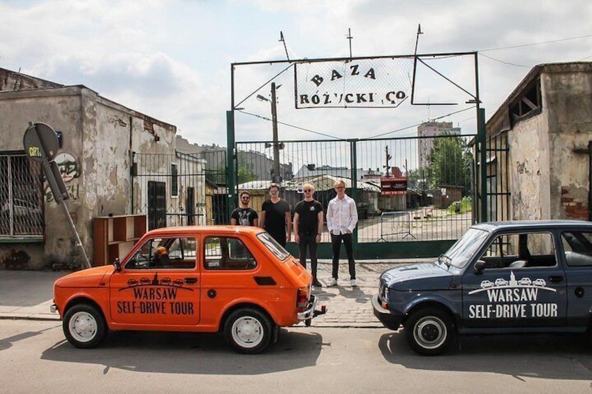Deux voitures anciennes, portant l’inscription « Visite guidée de Varsovie en voiture », sont garées devant un portail sur lequel on peut lire « Baza Rozwójki Co. ». 2
