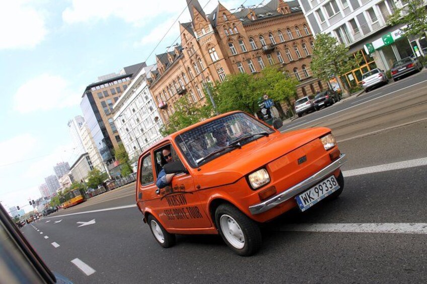A vintage orange car driving on a city street with historic buildings in the background. 1