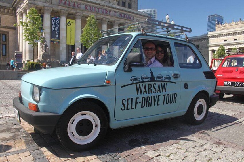 A light blue self-drive tour car with 'Warsaw Self-Drive Tour' written on the side parked on a cobblestone street. 4