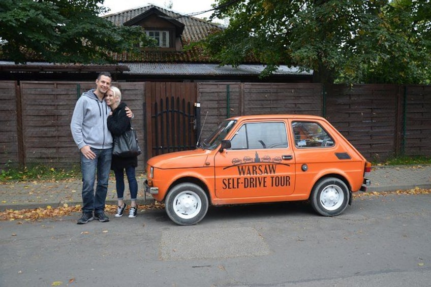 A vintage orange car with 'Warsaw Self-Drive Tour' written on it, parked on a street with a wooden fence and a house in the background. 2