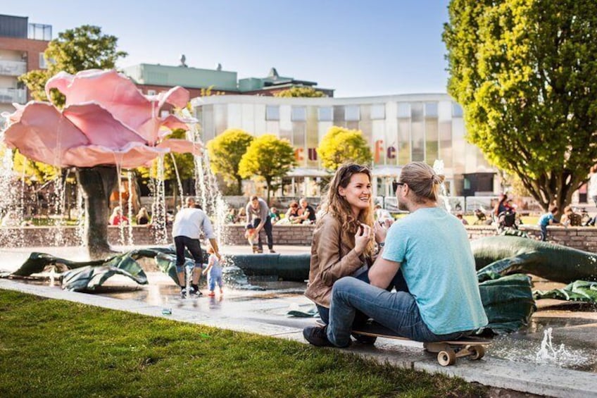 A couple sitting on a skateboard near a fountain with a large pink flower sculpture. 2