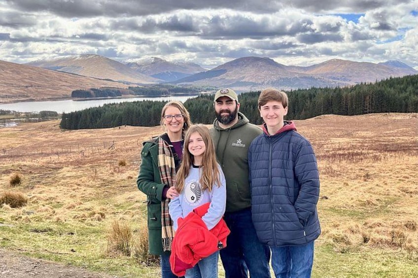 En familie på fire i et bjergrigt landskab med en sø og skov i baggrunden. 2