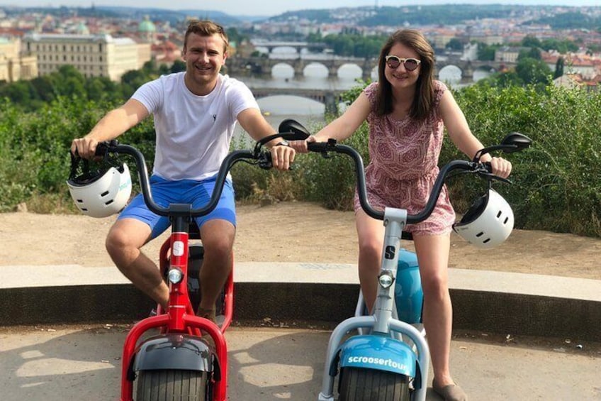 Två personer åker på elsparkcyklar på en asfalterad gångväg med en stadsbild i bakgrunden. 1