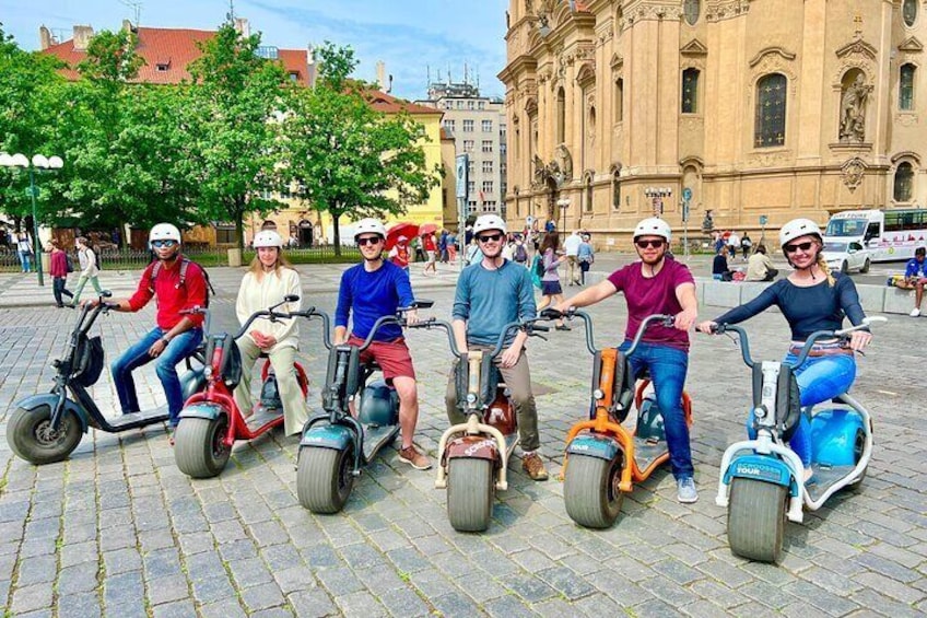 En grupp människor som åker elsparkcyklar på ett torg med kullerstenar, med historiska byggnader i bakgrunden. 2