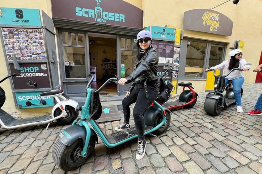 Two people riding electric scooters on a cobblestone street, with shops and signs in the background. 4
