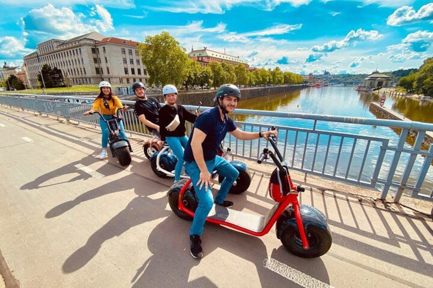 People riding electric scooters on a bridge with a river and buildings in the background. 4