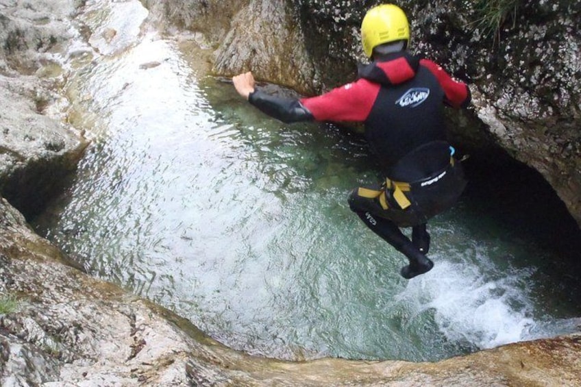 A person in a red and black wetsuit with a yellow helmet jumping into a rocky water body. 2