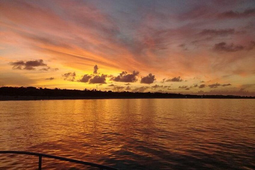 A sunset over a calm body of water with a silhouette of a distant shoreline and a railing in the foreground. 2