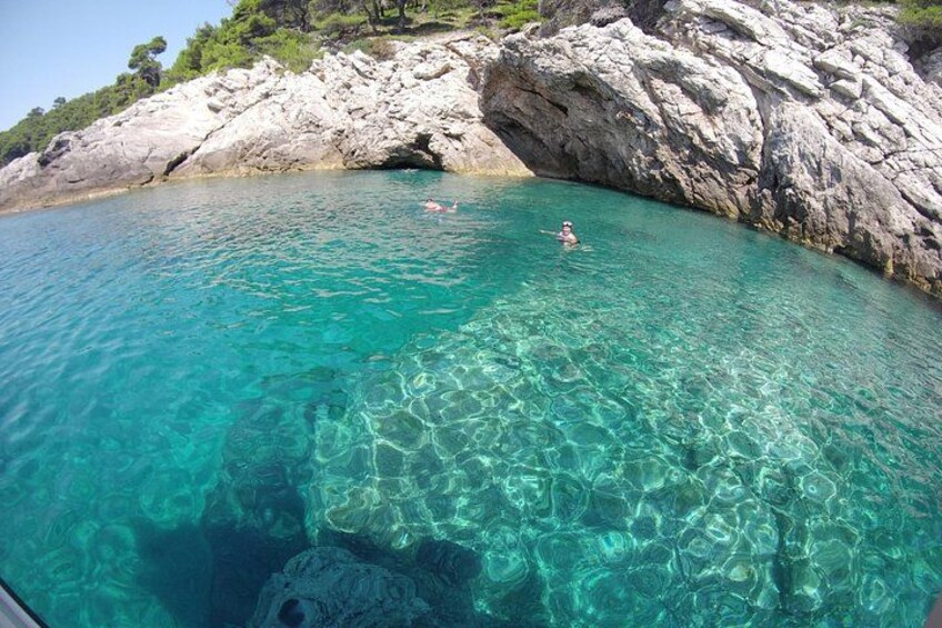 A clear turquoise sea with a rocky coastline, a person swimming, and another person paddling. 2