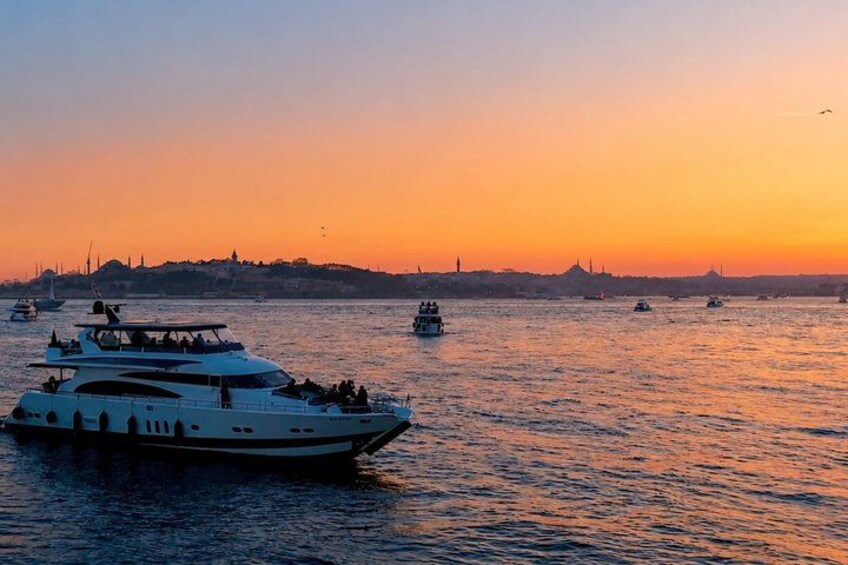Un coucher de soleil sur une étendue d’eau, avec plusieurs bateaux, dont un grand yacht, et une côte lointaine parsemée de bâtiments. 3