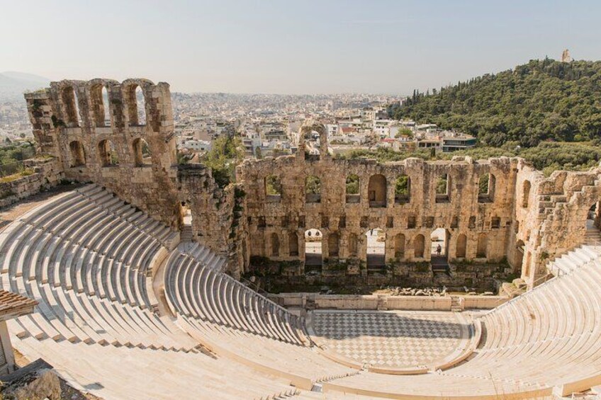 An ancient amphitheater with a partially intact stage and seating area, surrounded by ruins and a cityscape in the background. 2