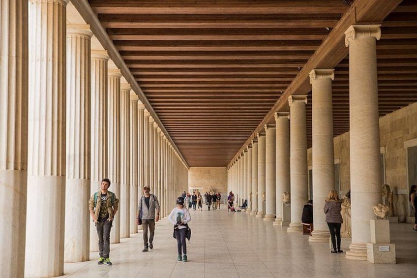 Un paseo techado con columnas, personas caminando y suelo de baldosas. 2