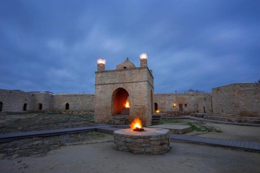 A stone building with a central archway and two chimneys, surrounded by walls and a fire pit in the foreground. 1