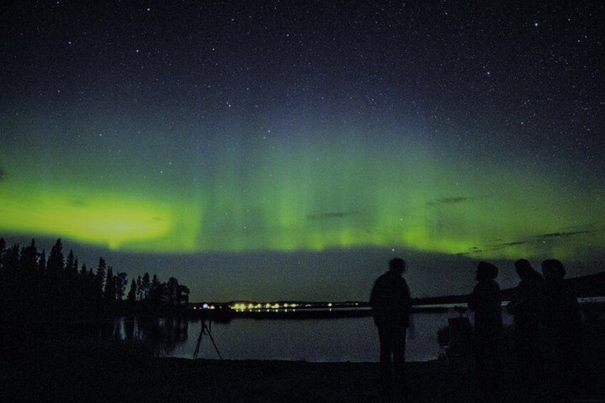 A night sky with the aurora borealis over a body of water, silhouettes of people observing, and a tripod with a camera. 5