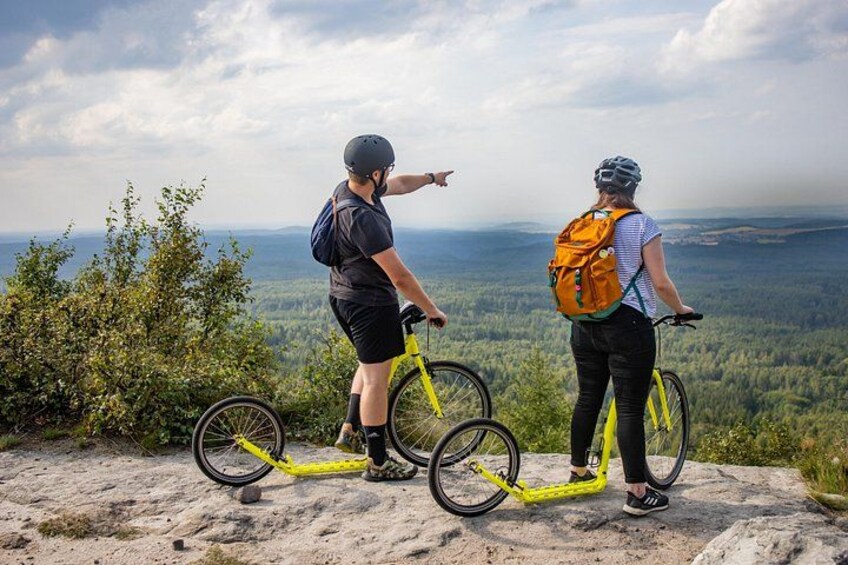 Two people on bicycles with backpacks, overlooking a forested landscape. 1
