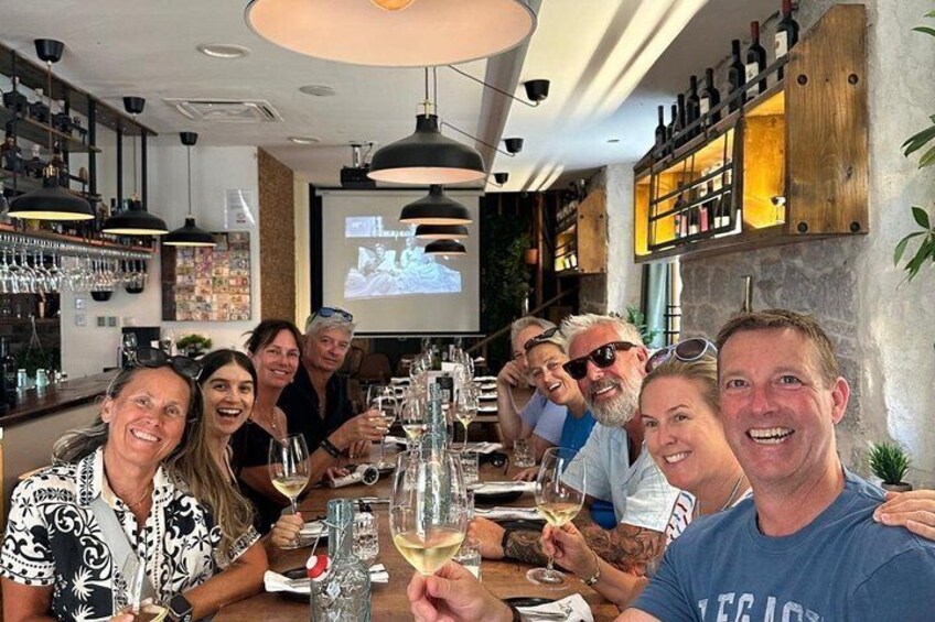 A group of people seated at a bar with wine glasses, a projector screen, and shelves of bottles. 4