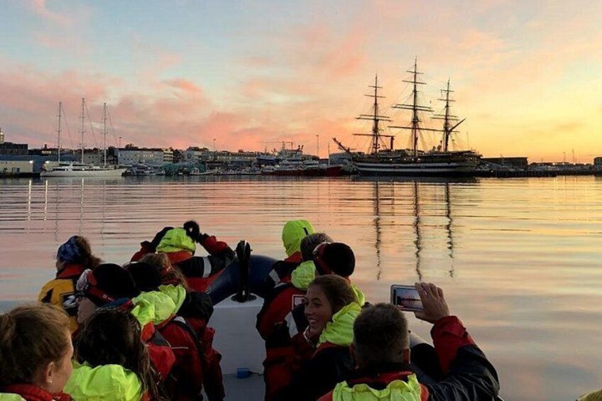 En grupp människor på en båt, ett stort segelfartyg och en lugn hamn vid solnedgången. 2