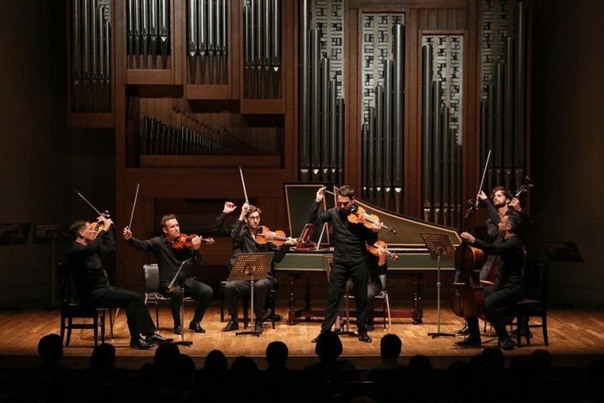 A classical music concert with musicians on stage, including violinists, a cellist, and a pianist, with a large organ in the background. 1