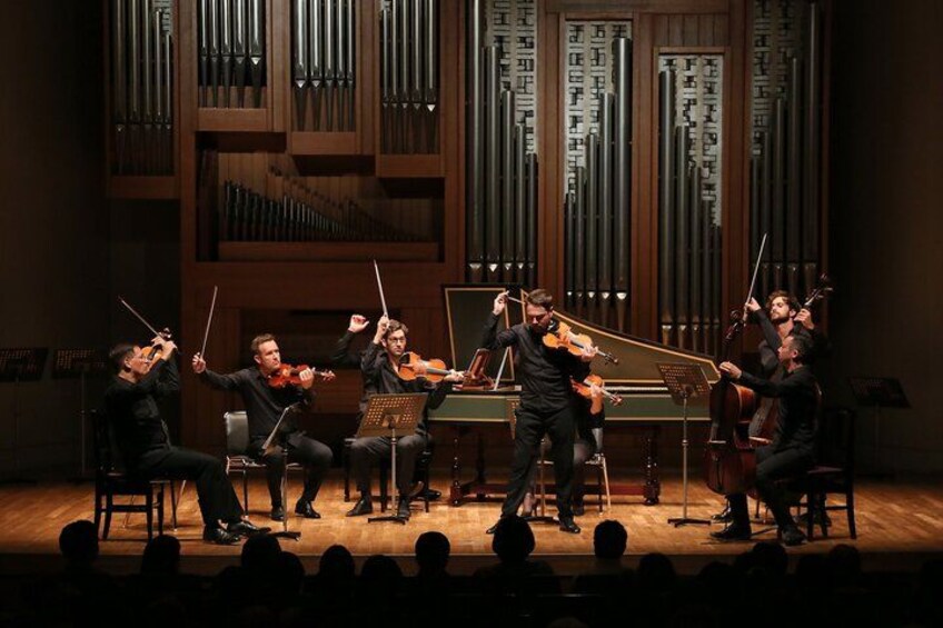 A classical orchestra performing on stage with musicians playing violins, cellos, and a piano. 1