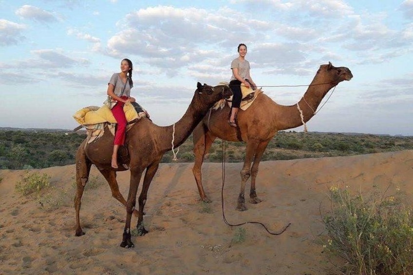 Two people riding camels on a sandy terrain with sparse vegetation. 3