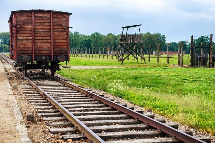 A rusty train car on tracks, a wooden structure resembling a watchtower, and a fenced area with trees in the background. 2