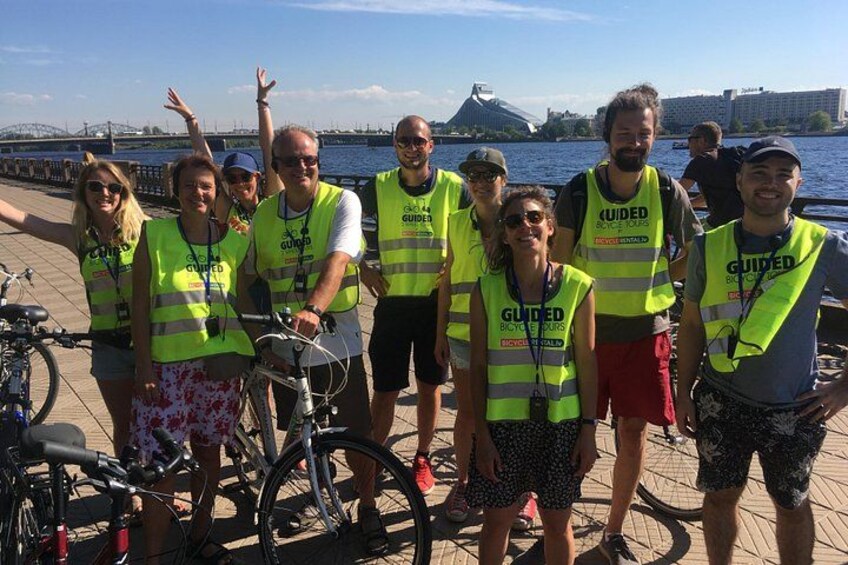Eine Gruppe von Menschen mit hochsichtbaren Westen und Fahrradbekleidung steht auf einer Uferpromenade. Im Hintergrund sind Fahrräder und eine Brücke zu sehen. 3