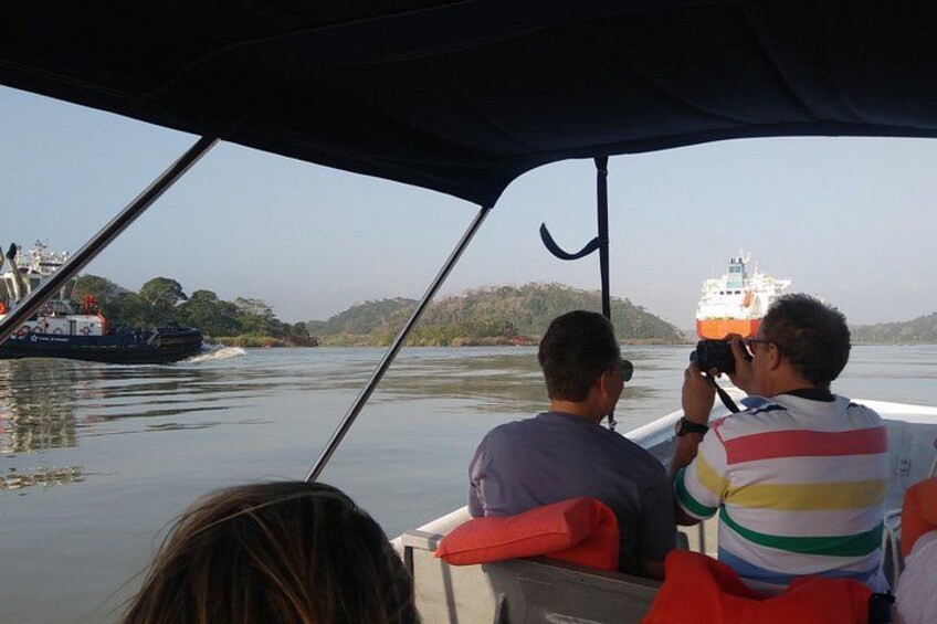 A boat with two people taking a photo, a large ship in the background, and a calm river. 2