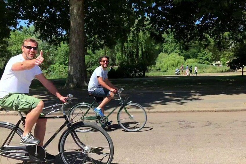 Two people riding bicycles on a paved path in a park with trees and a grassy area in the background. 4