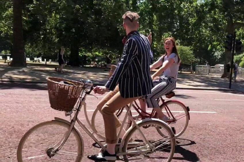 A man and a woman riding bicycles on a paved path in a park with trees and a stone wall in the background. 3