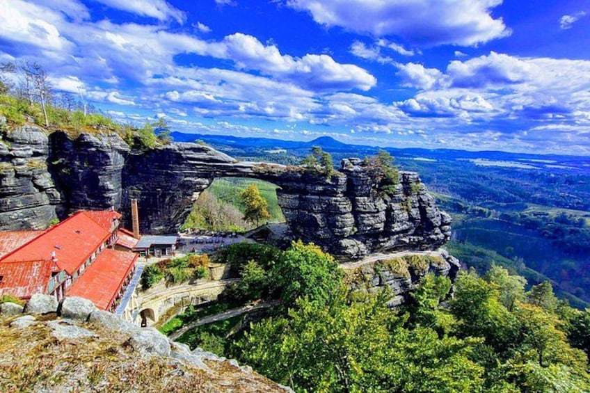 A rocky cliff with a red-roofed building, lush greenery, and a vast landscape under a partly cloudy sky. 1