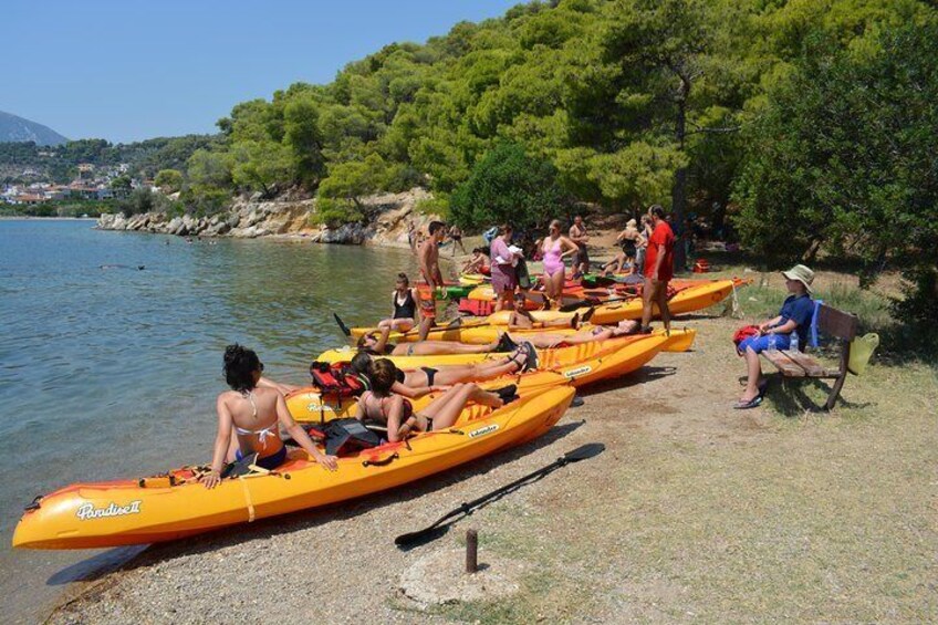 A group of people in orange kayaks on a riverbank, with trees and a clear sky in the background. 4