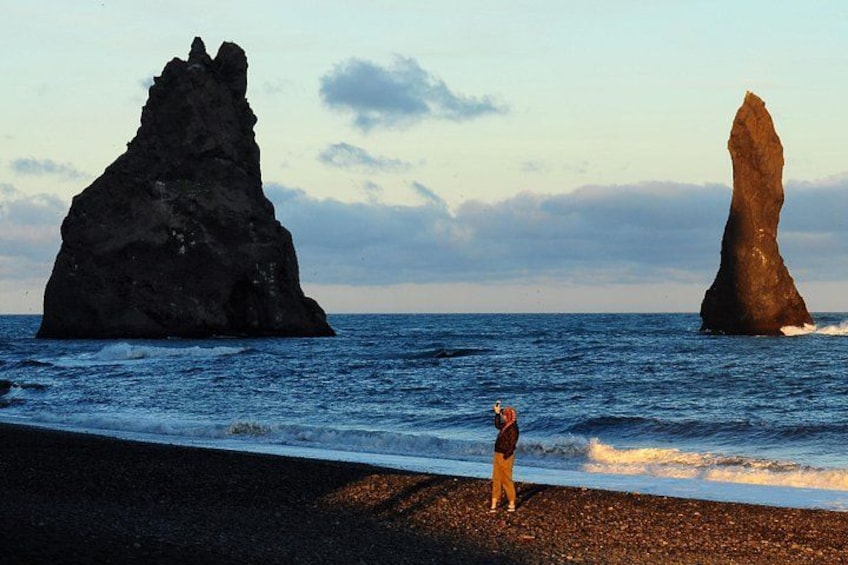 A rocky coastline with two prominent rock formations and a person standing on the pebbly beach. 1