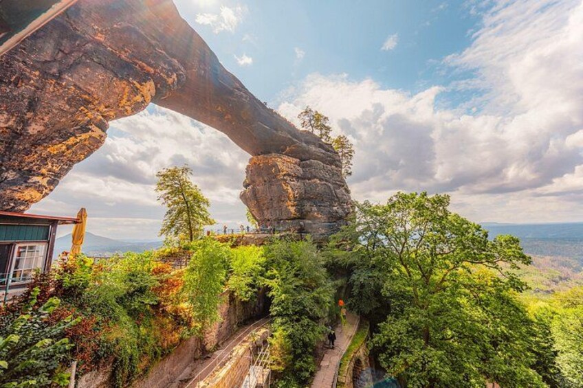 A natural rock formation arching over a trail with lush greenery and a distant view of the landscape. 3