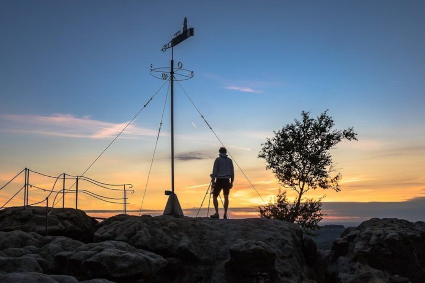 A person standing on a rocky outcrop with a cross at the top, overlooking a sunset. 4