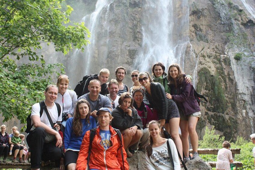 A group of people posing in front of a waterfall with a rocky cliff in the background. 1