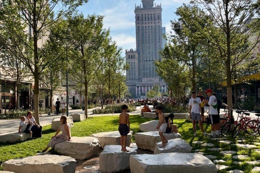 A park with people sitting on large rocks, trees, and a tall building in the background. 3