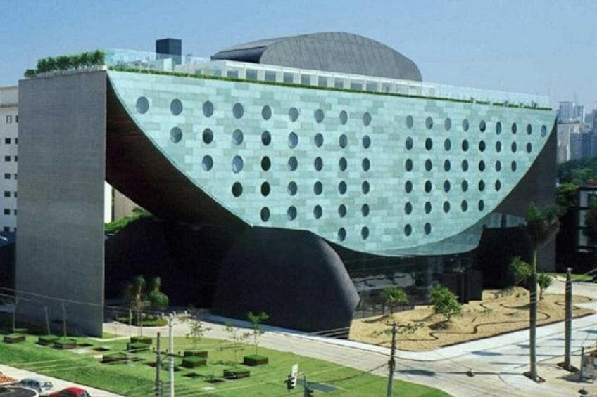 Ein modernes Gebäude mit einer gewölbten Glasfassade und kreisrunden Aussparungen, umgeben von einer begrünten Anlage mit Bäumen und blauem Himmel. 1