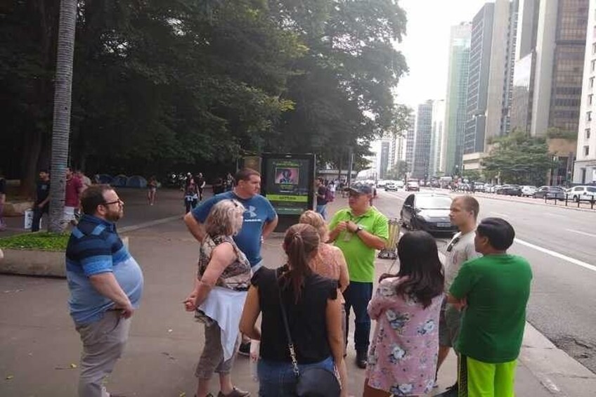 En grupp människor som står på en trottoar i ett tätbebyggt område, med höga byggnader i bakgrunden. 5